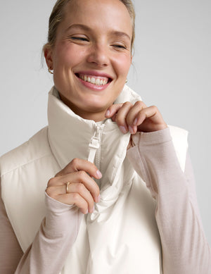 model wearing a white puffer vest over a long-sleeve shirt on a light gray background. 