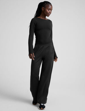 woman wearing a black long-sleeve boatneck bodysuit and black pants on a plain background. 
