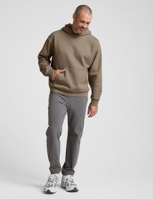 Man wearing a brown hoodie and gray pants on a plain background. 