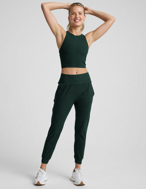 woman wearing dark green cropped tank and jogger pants on a plain background. 
