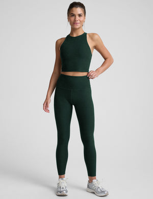 woman wearing a green high-neck racerback cropped tank and leggings on a plain background. 