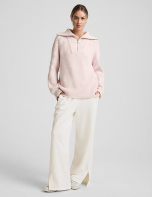 Woman wearing a light pink quarter zip sweater and white fleece pants against a plain background. 